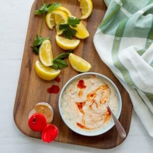 Wooden tray topped with a light blue bowl of Cajun tartar sauce with hot sauce on top, a bottle of hot sauce on the side, as well as lemon wedges, parsley leaves, and a kitchen towel.