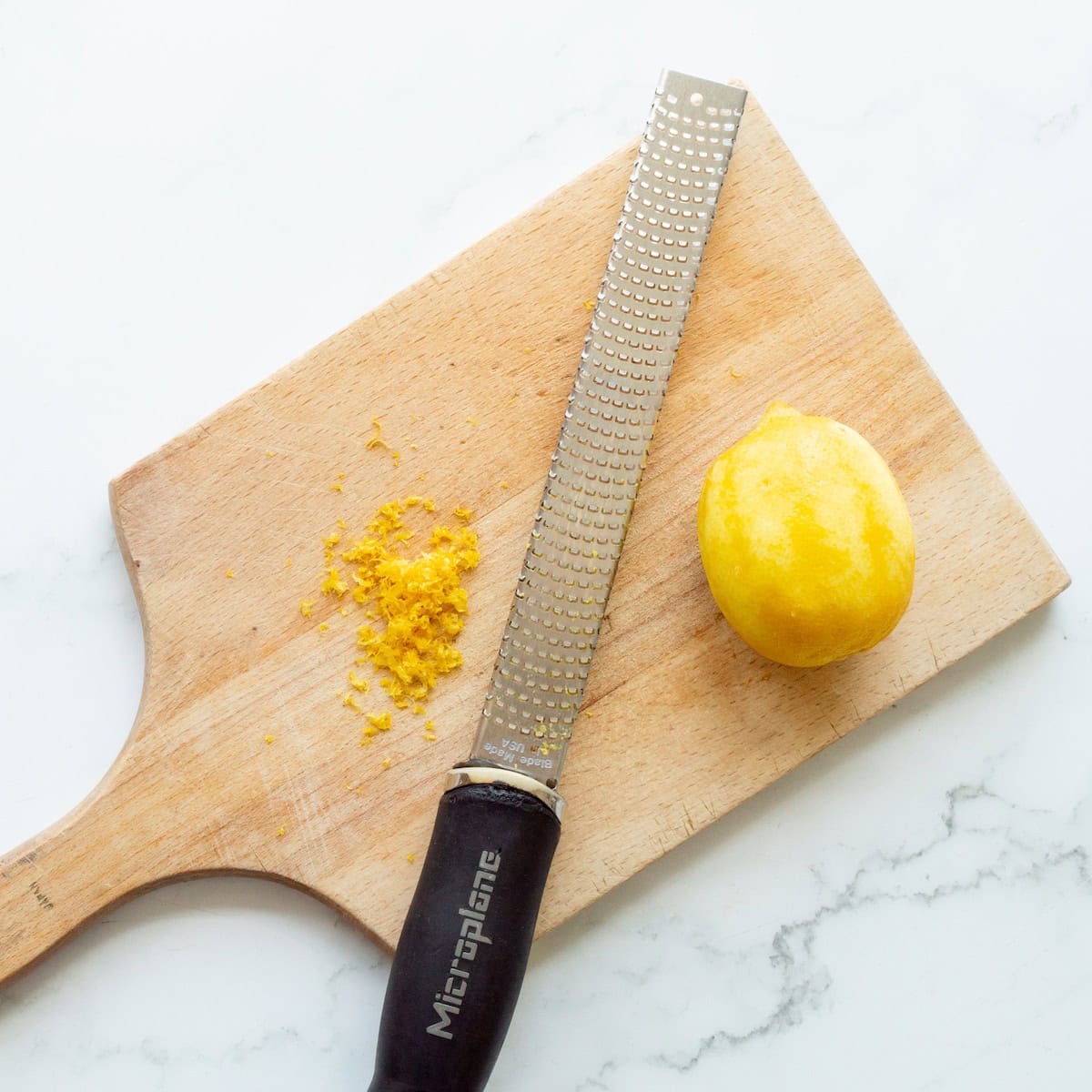 A zested lemon on a wooden cutting board with zest to the side and a Microplane.