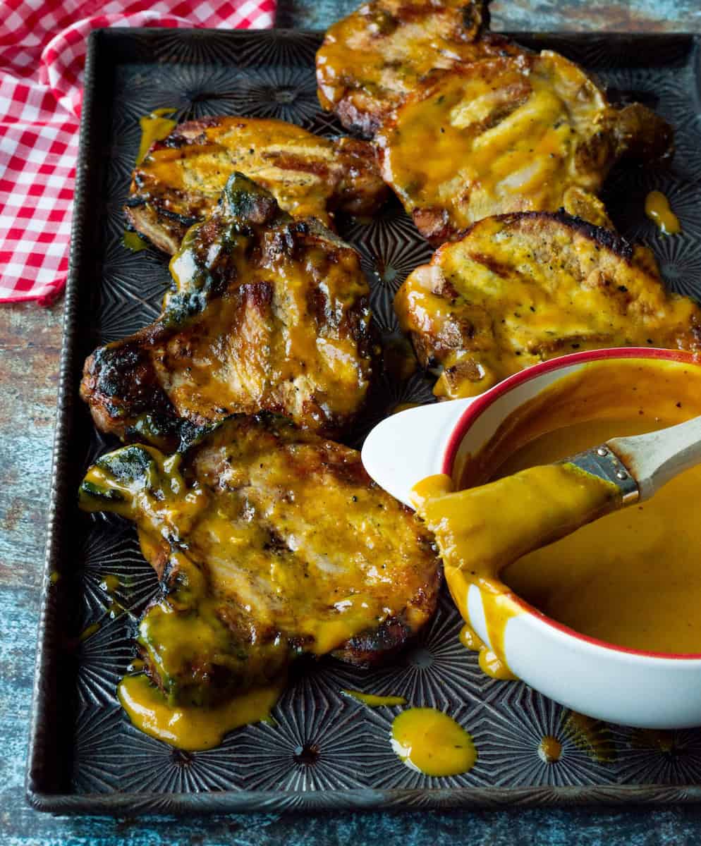 A metal pan of grilled pork chops brushed with Carolina Gold BBQ Sauce alongside a white bowl of sauce with a brush on top.