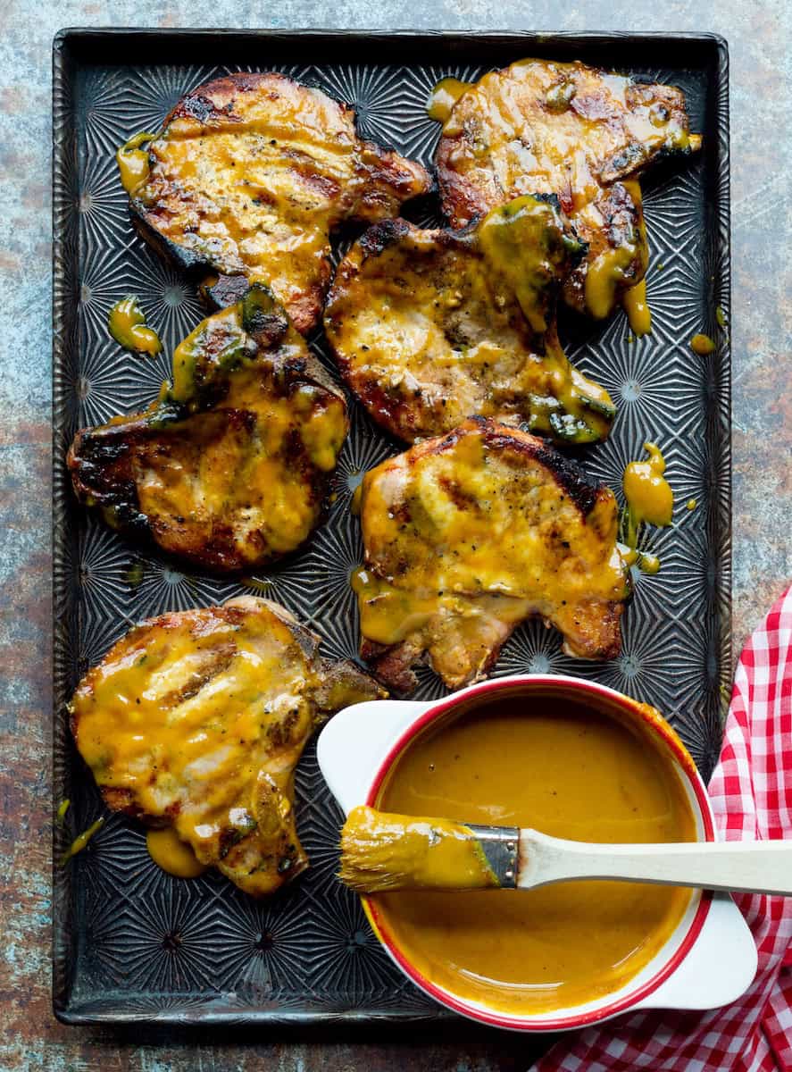 A metal pan of grilled pork chops brushed with Carolina Gold BBQ Sauce alongside a white bowl of sauce with a brush on top and a red checked cloth.