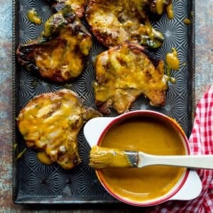 A metal pan of grilled pork chops brushed with Carolina Gold BBQ Sauce alongside a white bowl of sauce with a brush on top and a red checked cloth.