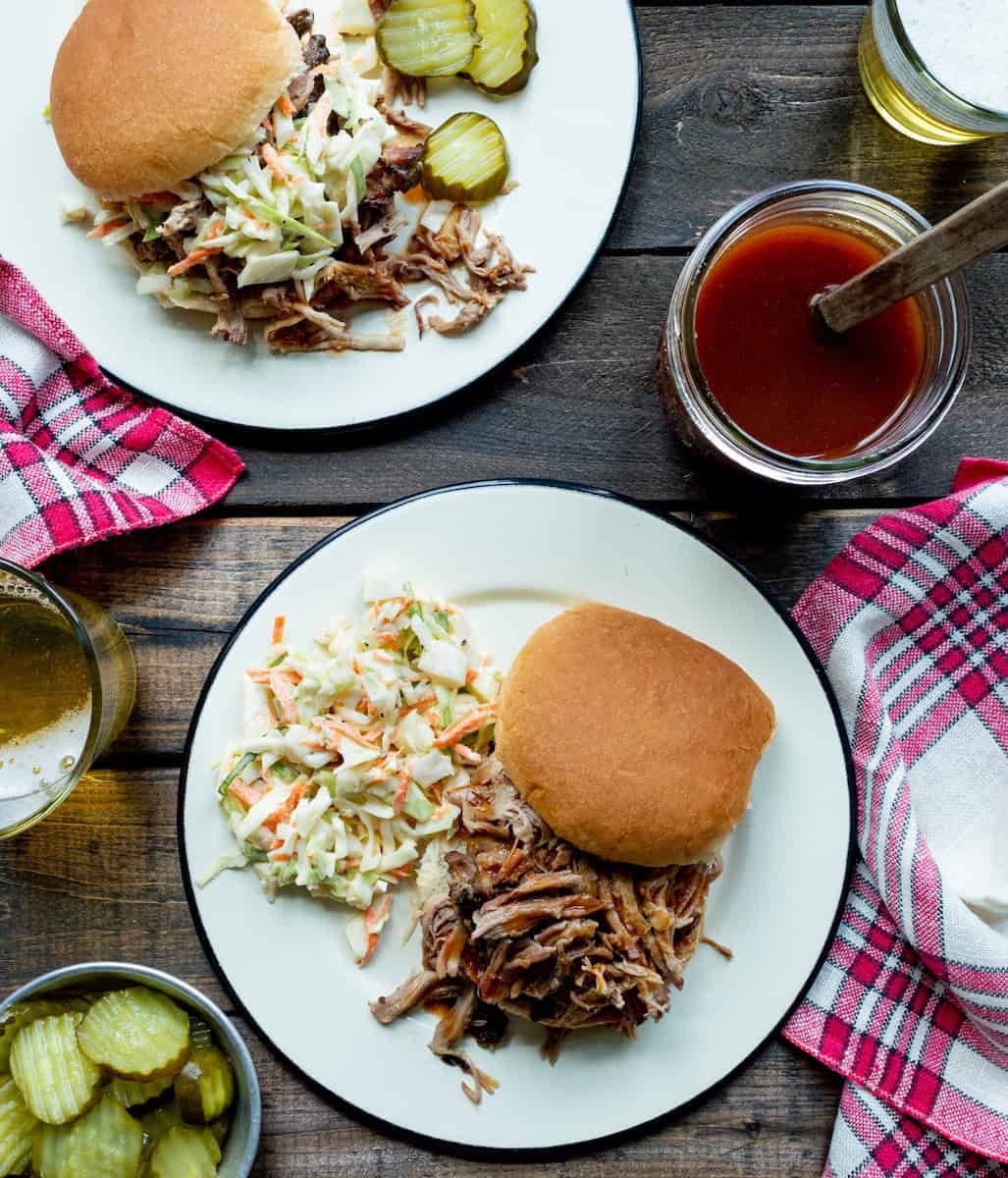 Two plates topped with pulled pork sandwiches and coleslaw, a jar of Western NC BBQ Sauce, a bowl of pickles, and some red checked napkins.