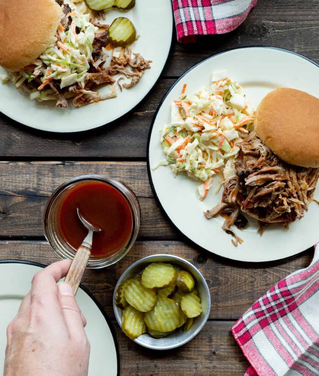 Two plates topped with pulled pork sandwiches and coleslaw, a jar of Western NC BBQ Sauce with a hand spooning some out, a bowl of pickles, and some red checked napkins.