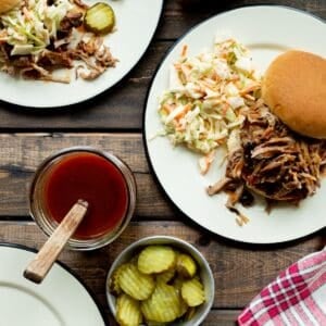 Two plates topped with pulled pork sandwiches and coleslaw, a jar of Western NC BBQ Sauce, a bowl of pickles, and some red checked napkins.
