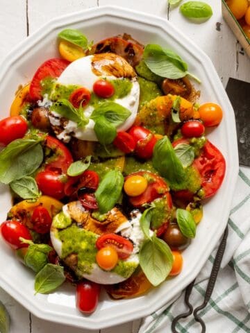 White platter of Burrata Caprese Salad with a metal spatula and green checked cloth on the side.