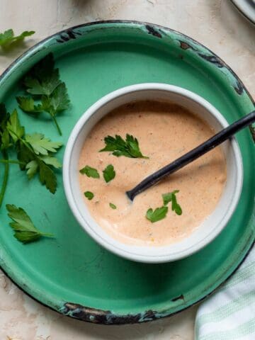 Remoulade sauce in a white bowl on a green enamel plate with parsley leaves on the side, a metal spoon in the bowl, a green striped cloth to the side, a stack of plates, and glasses of water.