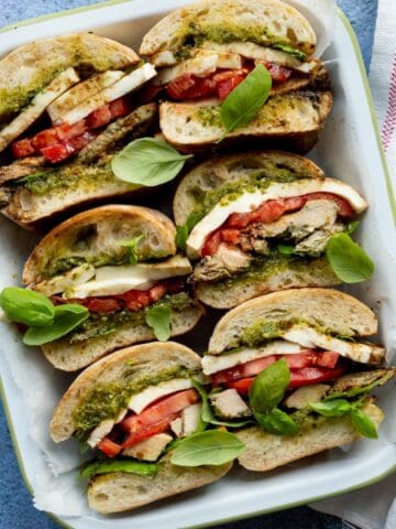 Trader Joe's pesto chicken caprese sandwiches cut in half and lined up in a white enamel pan beside a red lined white napkin.