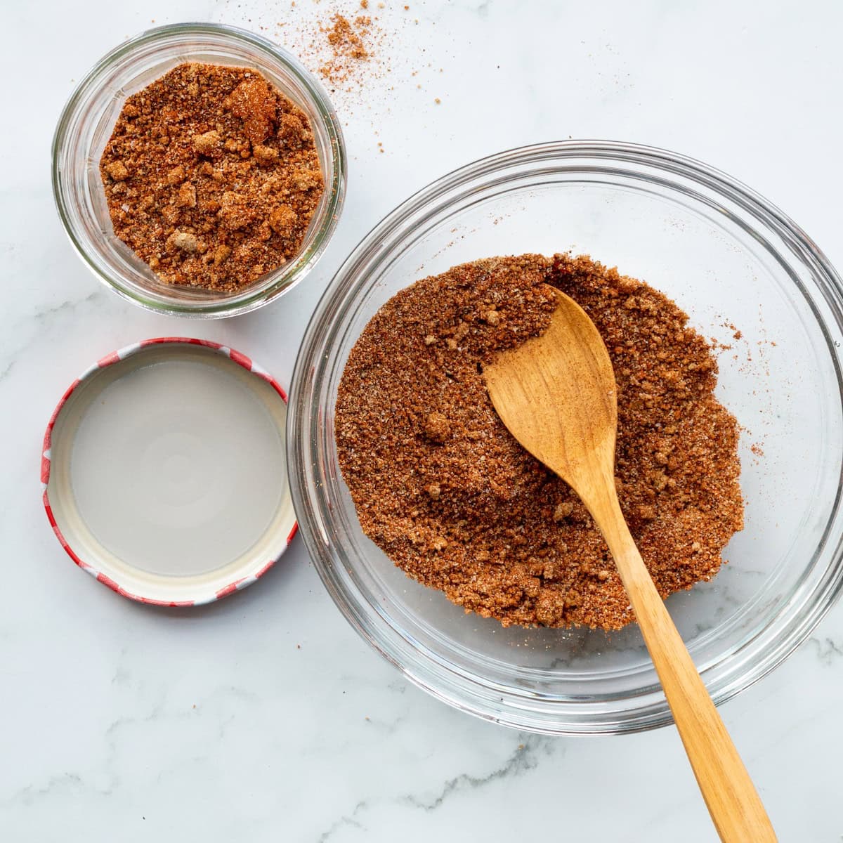 Desert Heat Dry Rub in a glass bowl with a wooden spoon with some of the rub in a Bonne Maman jar off to the side.