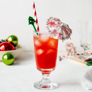A clear glass filled with the Mele Kalikimaka Christmas rum tiki drink and garnished with a paper umbrella, Christmas tree swizzle stick, and red striped straw.