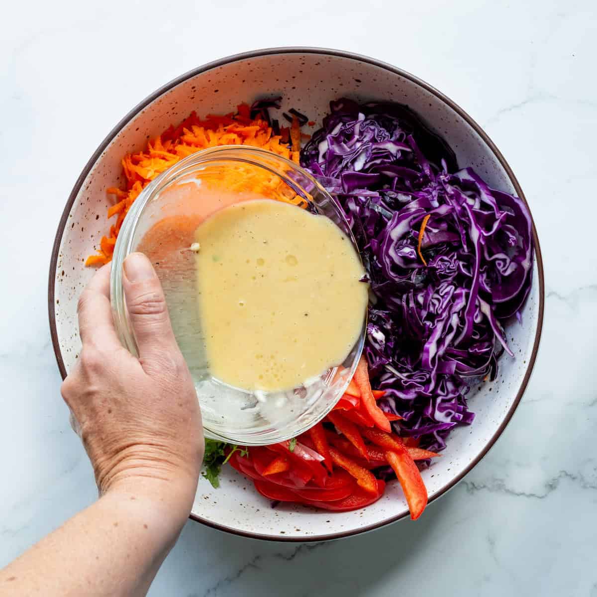 Pouring dressing over purple cabbage, red bell peppers, and carrots in a white bowl.