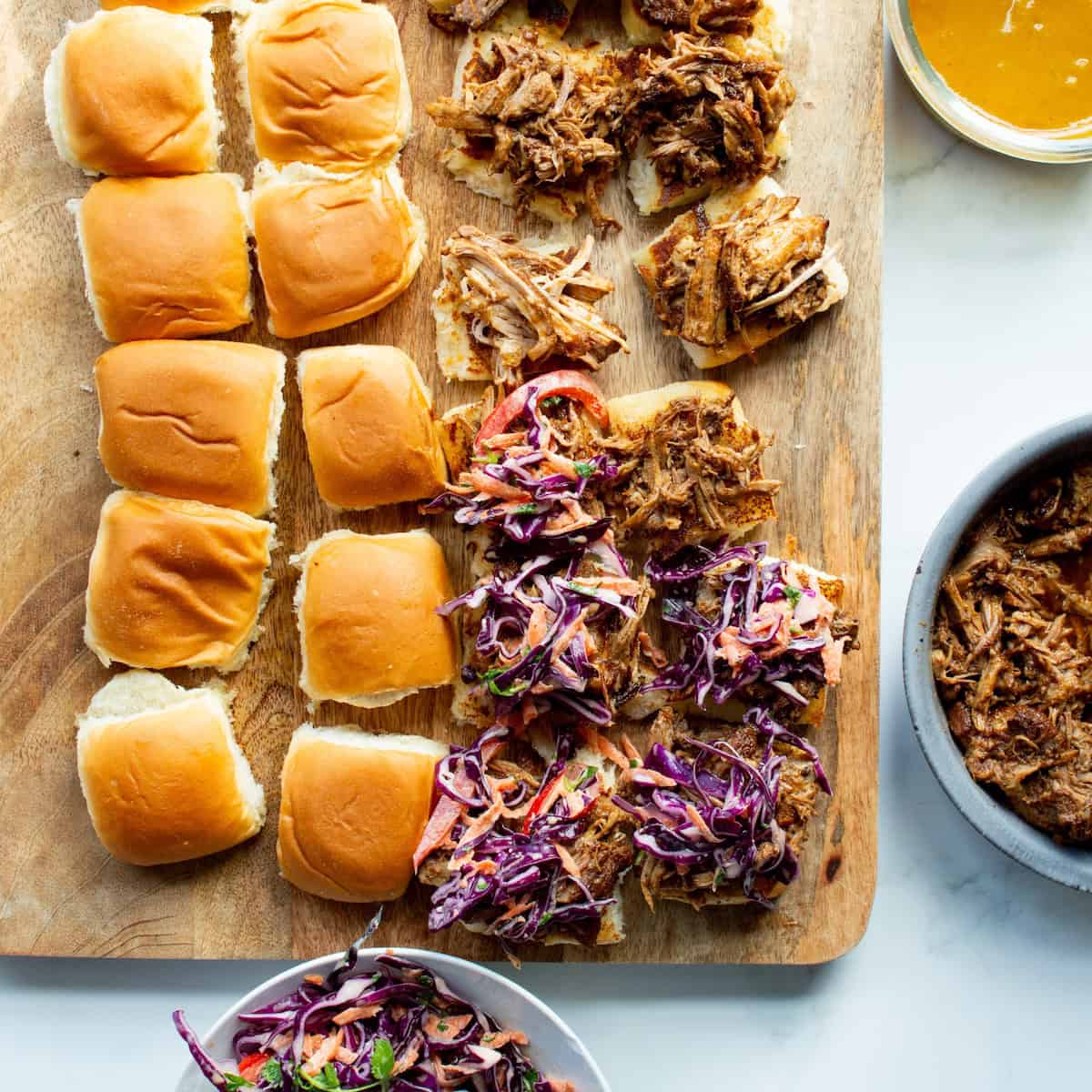 Assembling BBQ Pulled Pork Sliders on a wooden board with the filling ingredients surrounding.