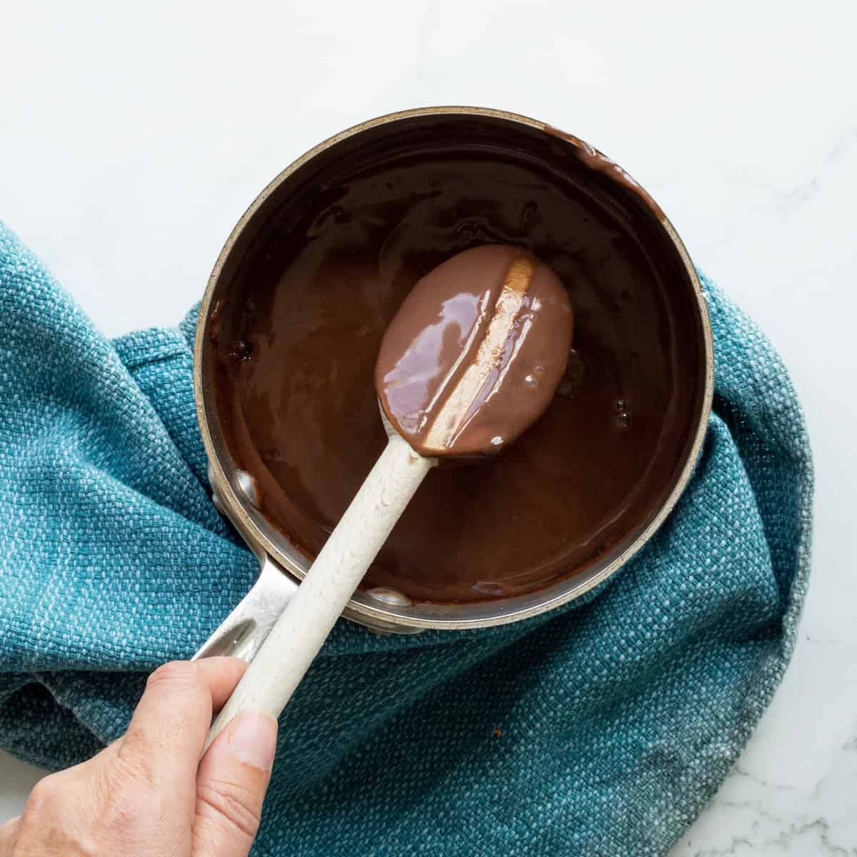 Cooked chocolate pudding in a small saucepan with a wooden spoon and a line drawn through pudding on the spoon to indicate doneness.
