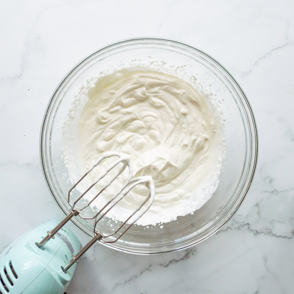 Whipped cream in a clear bowl with an aqua hand mixer off to the side.