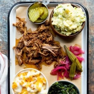 Enamel tray topped with Dutch oven pulled pork, bowls of macaroni and cheese, collard greens, dill pickle chips, and potato salad, and a pile of pickled red onions and okra beside a red-striped white napkin.