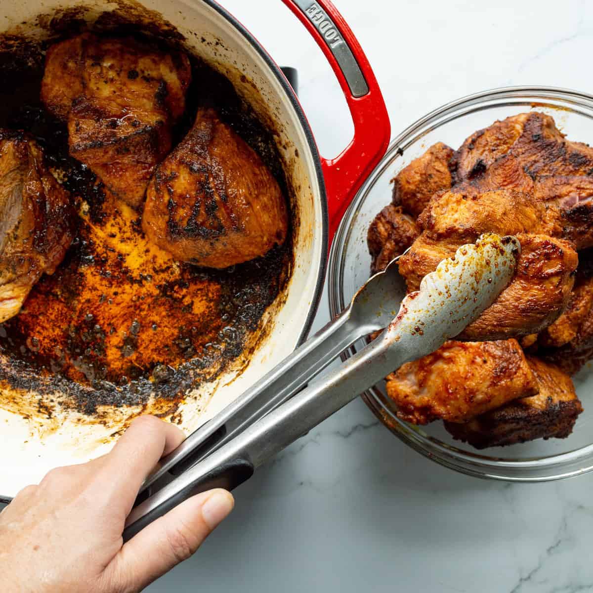 Removing seared pieces of pork from a red handled Dutch oven to a glass bowl with tongs.