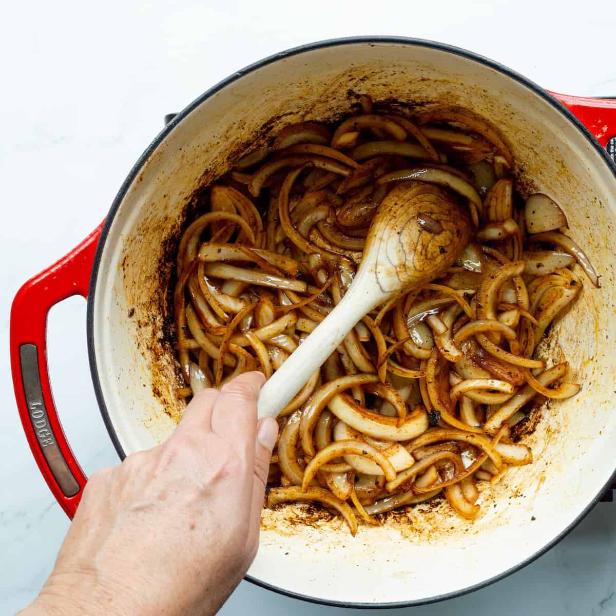 Sautéing onions with a wooden spoon in pork drippings in a red handled Dutch oven.
