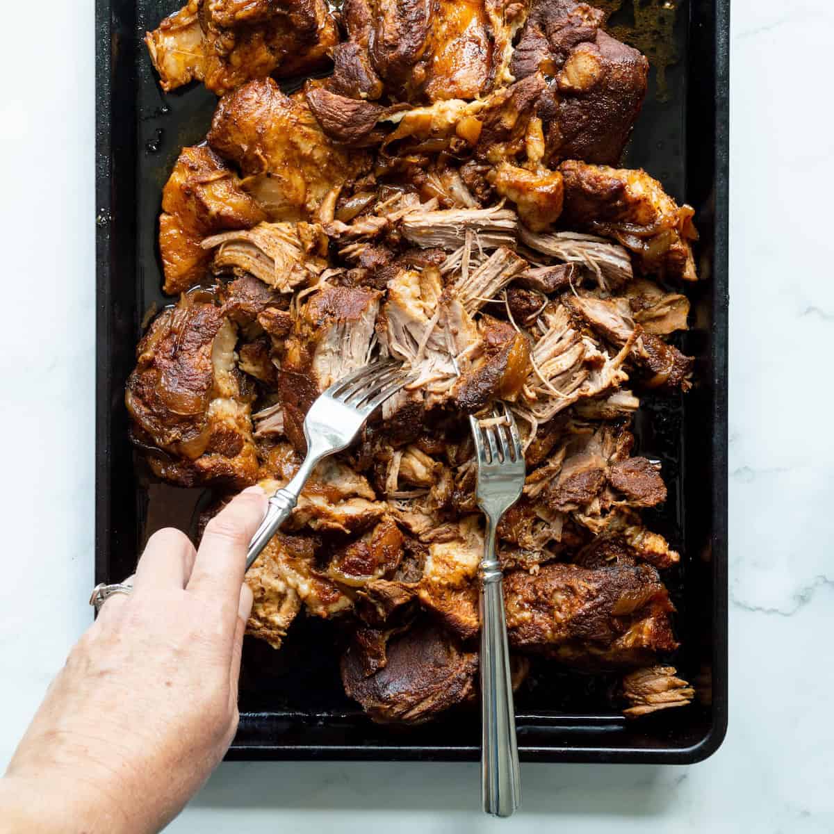 Shredding slow-cooked pork with two forks on a dark metal baking pan.