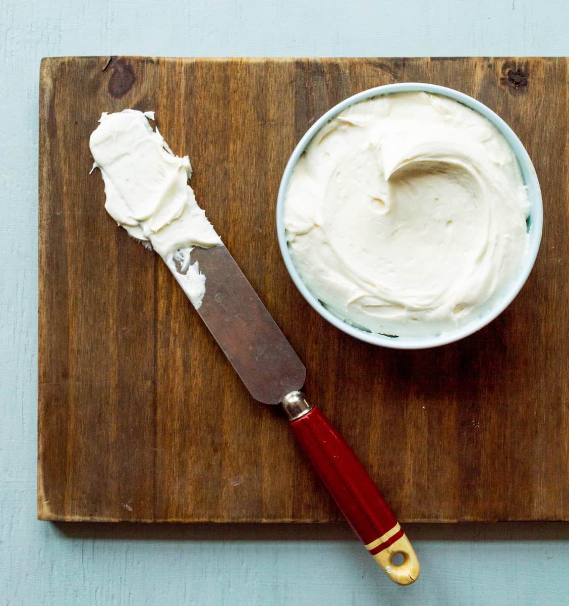 A wooden board topped with a small light blue bowl of cream cheese frosting with a red handled vintage spatula dipped in frosting on the side.