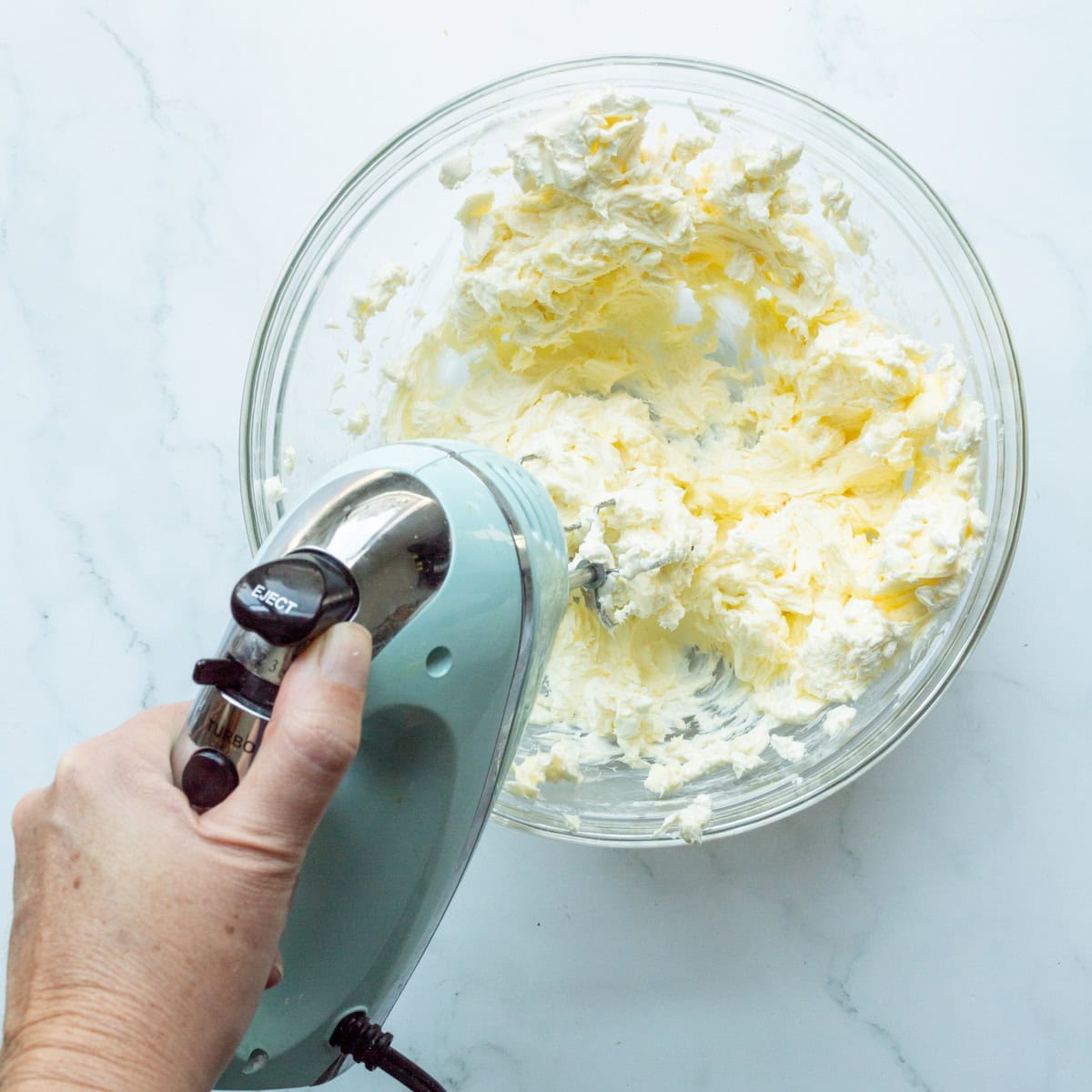 Beating cream cheese and butter in a glass bowl with an aqua hand mixer.