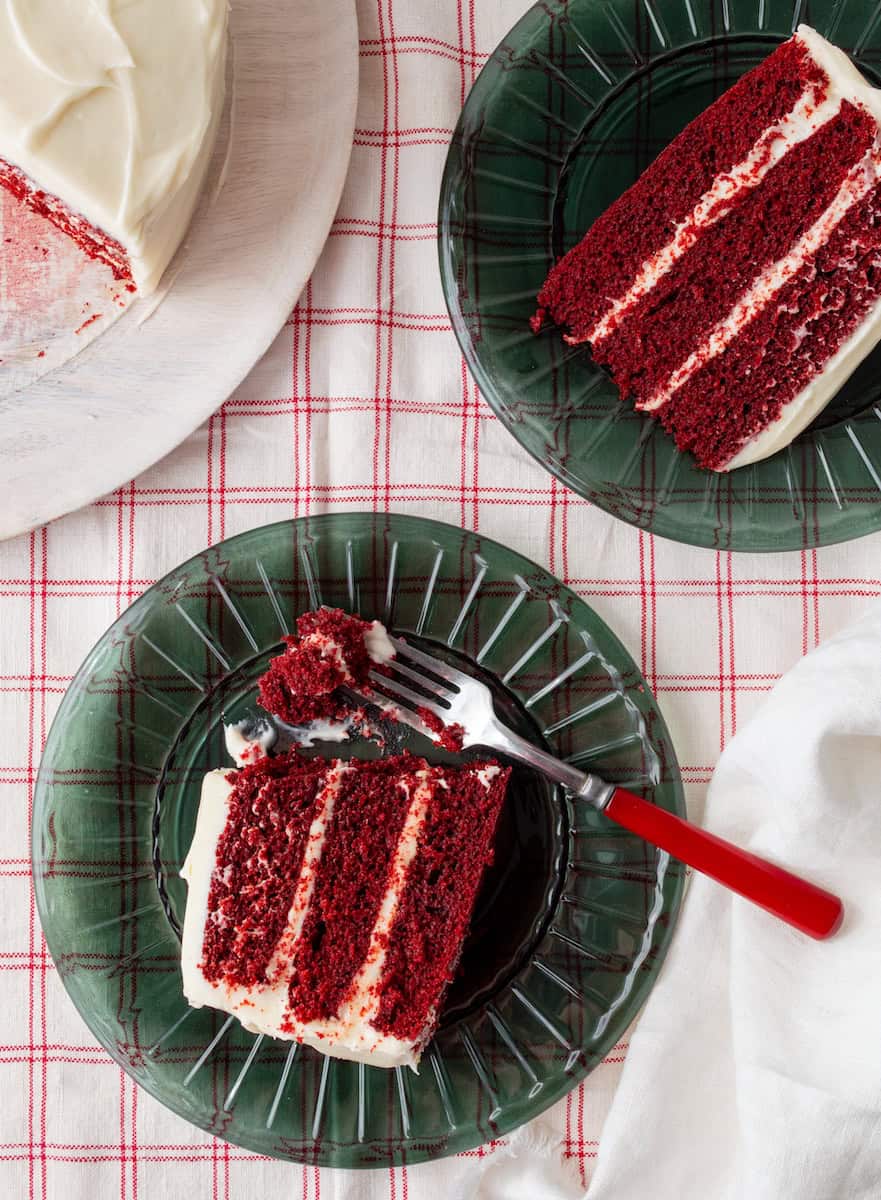 Slices of red velvet cake on green plates with a bite on a red handled fork with a platter of cake off to the side.
