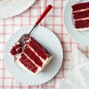 Slice of red velvet cake on a white plate with a bite on a red handled fork with another slice on a plate.