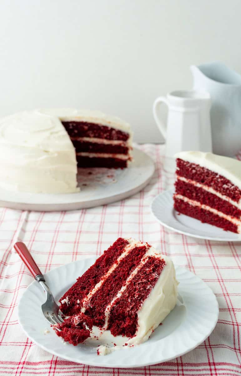 Slice of red velvet cake on a white plate with a bite on a red handled fork with another slice on a plate and platter of cake in the background.