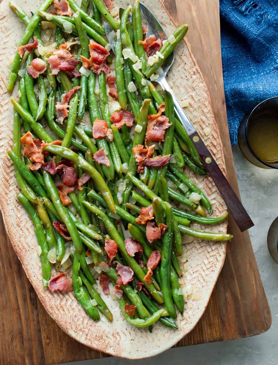 Wooden board topped with an oval pottery platter with Southern Green Beans with Onions and Bacon with a wooden handled serving fork, a glass of water, and a blue cloth on the side.