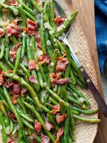 Wooden board topped with an oval pottery platter with Southern Green Beans with Onions and Bacon with a wooden handled serving fork, a glass of water, and a blue cloth on the side.