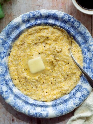 Antique blue bowl filled with yellow Creamy Southern grits topped with a melted butter pat, with a spoon, cream colored napkin, cup of black coffee, and green salt and pepper shakers.