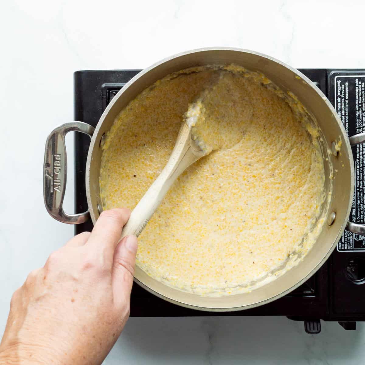 Stirring a pot of creamy yellow grits with a wooden spoon in a medium saucepan.