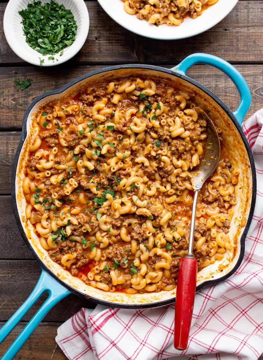 Teal handled skillet filled with Hamburger Helper without Milk with a red handled spoon, red checkered cloth, bowl of parsley, and serving bowl of the hamburger helper.