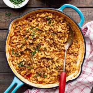 Teal handled skillet filled with Hamburger Helper without Milk with a red handled spoon, red checkered cloth and bowl of parsley.