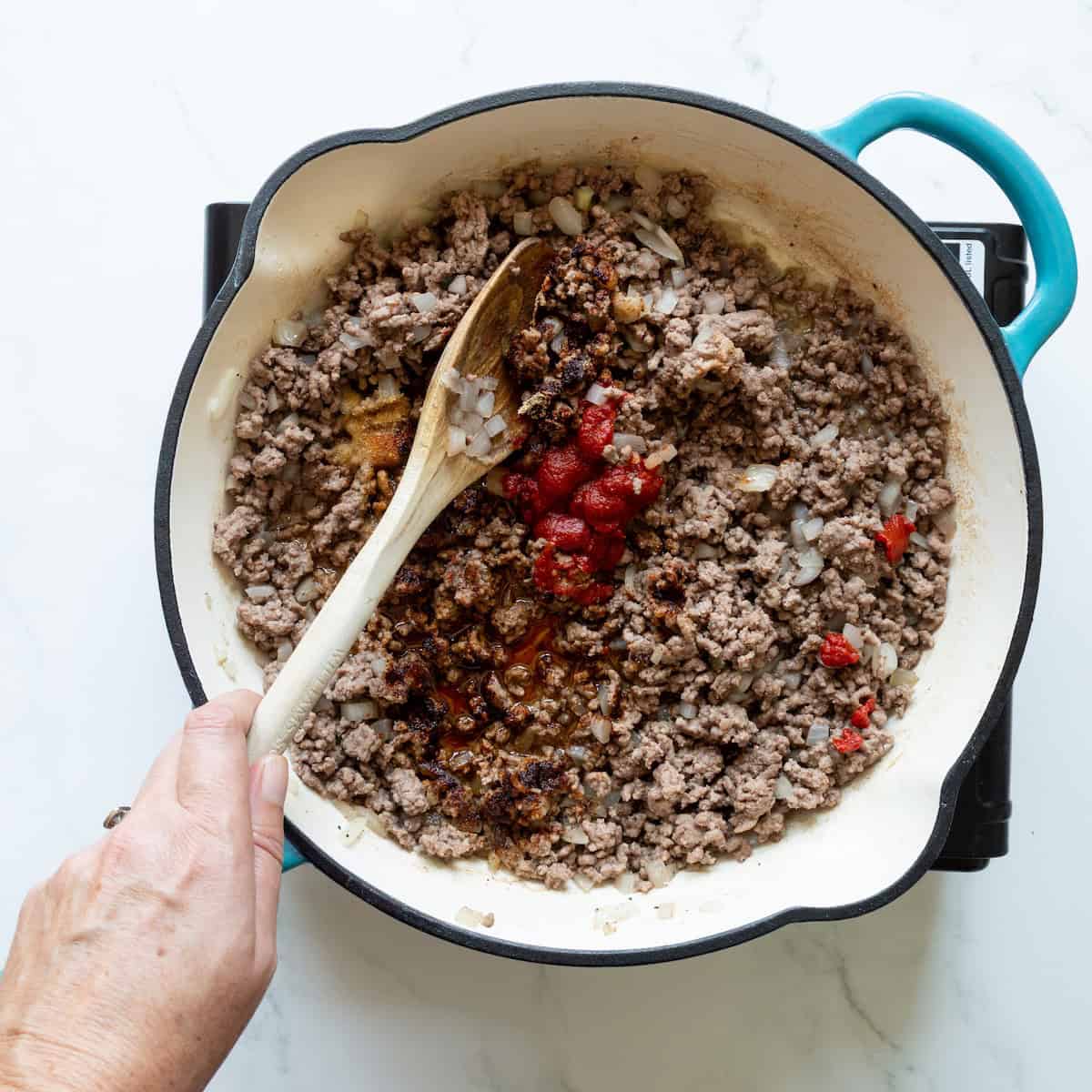 Adding tomato paste and seasoning to browned ground beef and onion in a teal-handled skillet.