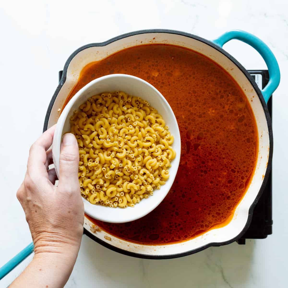 Adding uncooked macaroni to a teal-handled skillet filled with a red sauce.