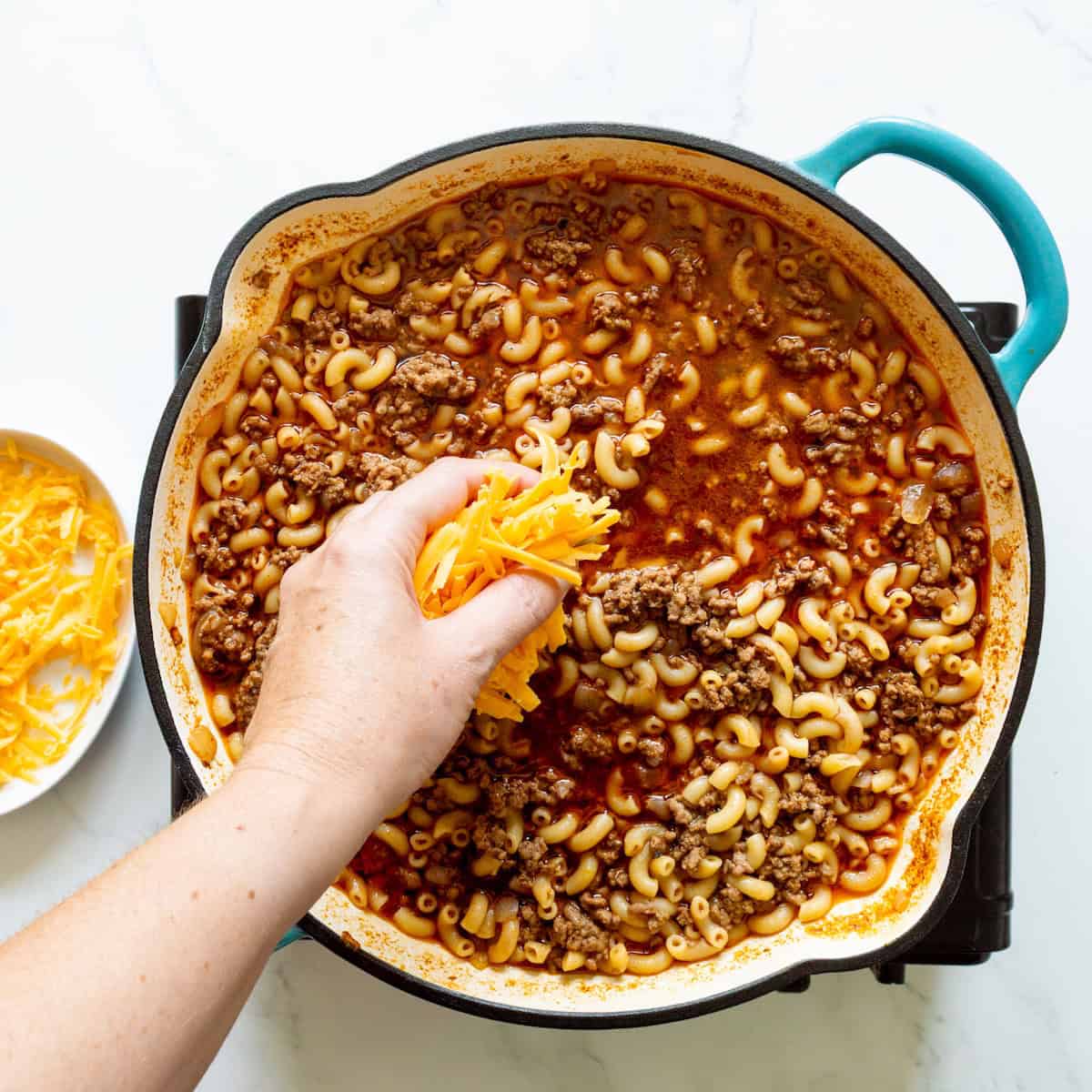 Adding cheese to the cooked homemade hamburger helper in a teal handled skillet with a bowl of cheese off to the side.