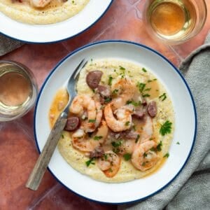 Two blue rimmed white bowls filled with Magnolia's Charleston Shrimp and Grits with two glasses of white wine, a wooden handled fork, and a tan cloth.