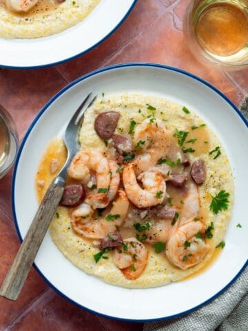 Two blue rimmed white bowls filled with Magnolia's Charleston Shrimp and Grits with two glasses of white wine, a wooden handled fork, and a tan cloth.