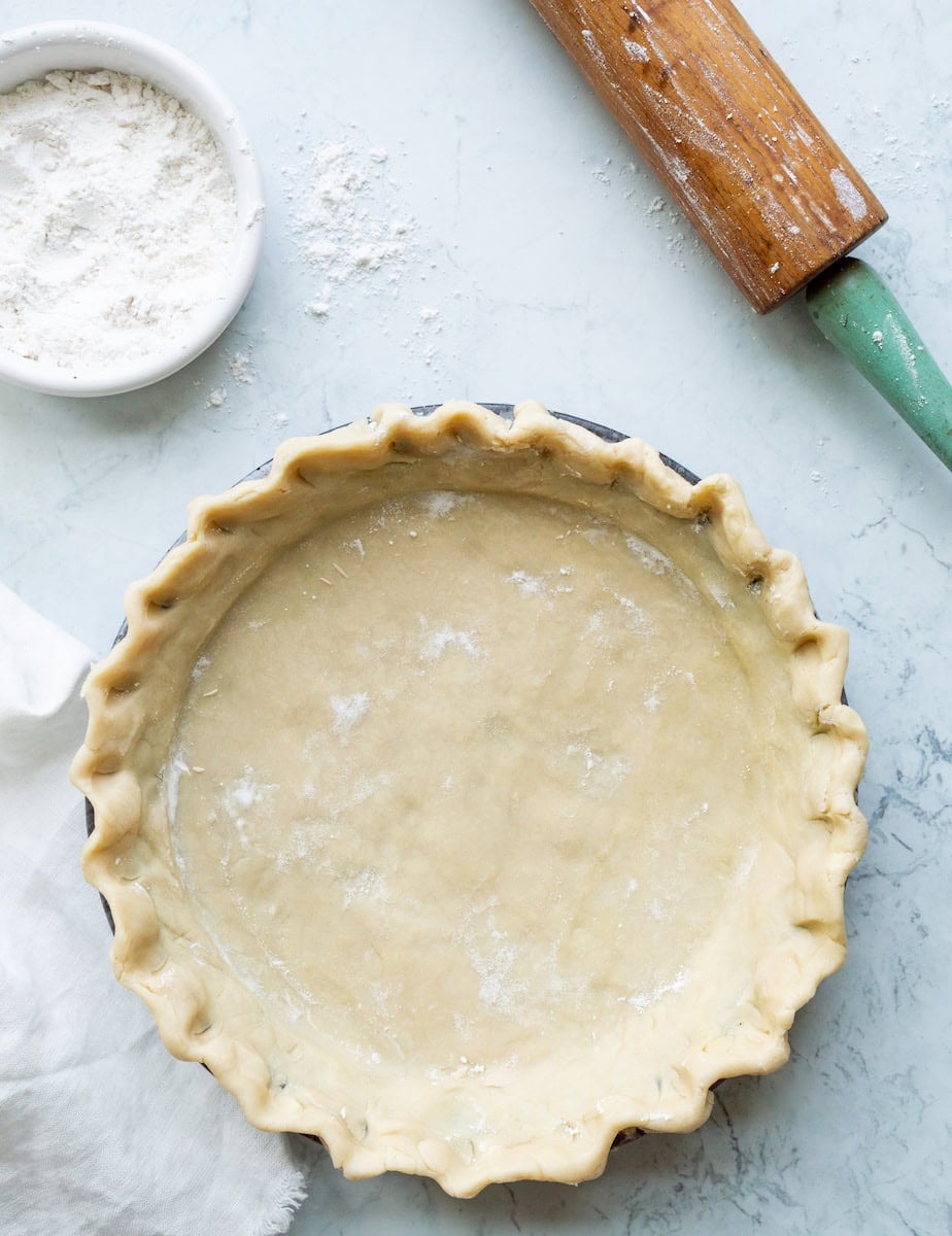 Crimped uncooked all butter pie crust in a pie tin with a white cloth, white bowl of flour, and green handled rolling pin off to the side.