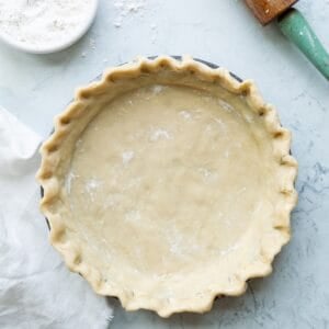 Crimped uncooked all butter pie crust in a pie tin with a white cloth, white bowl of flour, and green handled rolling pin off to the side.