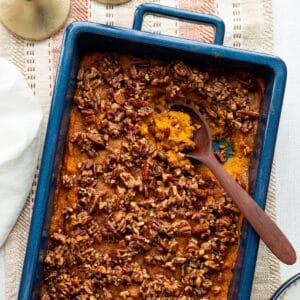 Sweet potato casserole with pecan streusel topping in a blue casserole dish with a wooden spoon and small blue rimmed china plates.