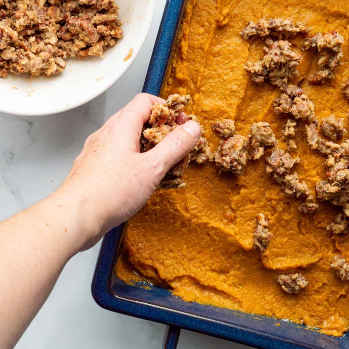 Sprinkling streusel on top of sweet potato casserole in a blue casserole dish.