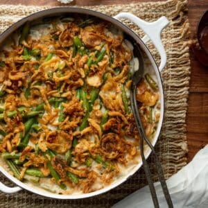 Green Bean Casserole with French's onions in a white round casserole dish on top of a textured cloth with salt and pepper shakers and a metal serving spoon.