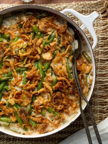 Green Bean Casserole with French's onions in a white round casserole dish on top of a textured cloth with salt and pepper shakers and a metal serving spoon.