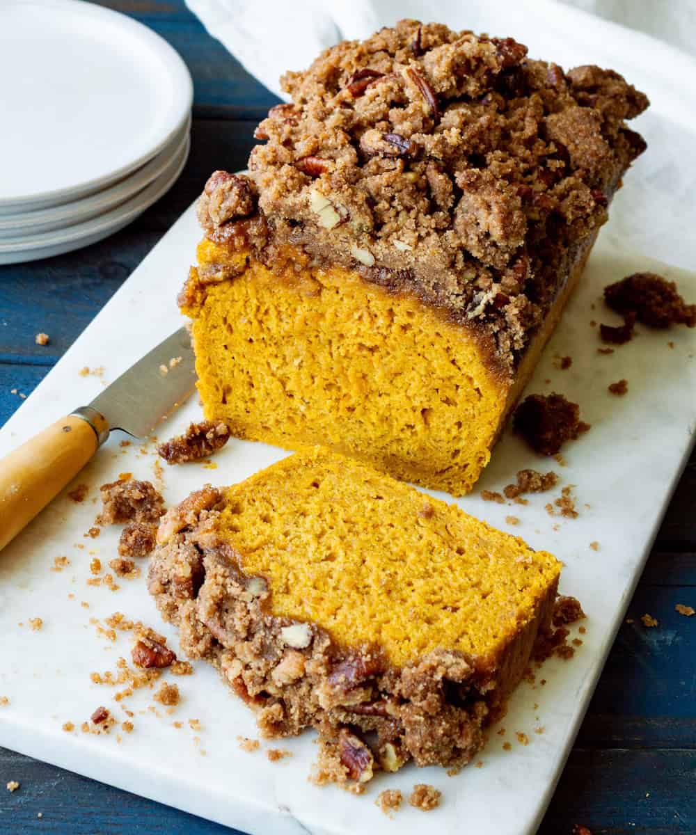 Sliced streusel topped pumpkin bread on a white marble board with a paring knife on the side and stack of white plates in the background.