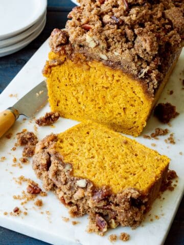 Sliced streusel topped pumpkin bread on a white marble board with a paring knife on the side and stack of white plates in the background.