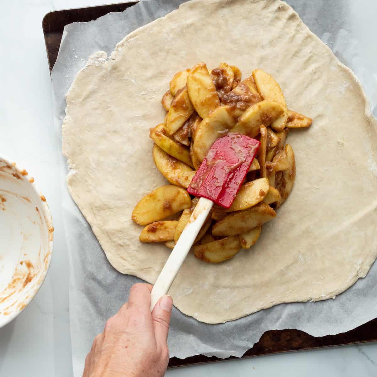 Spooning apple filling with a red spatula into the center of a round of pie dough on a parchment lined baking sheet.