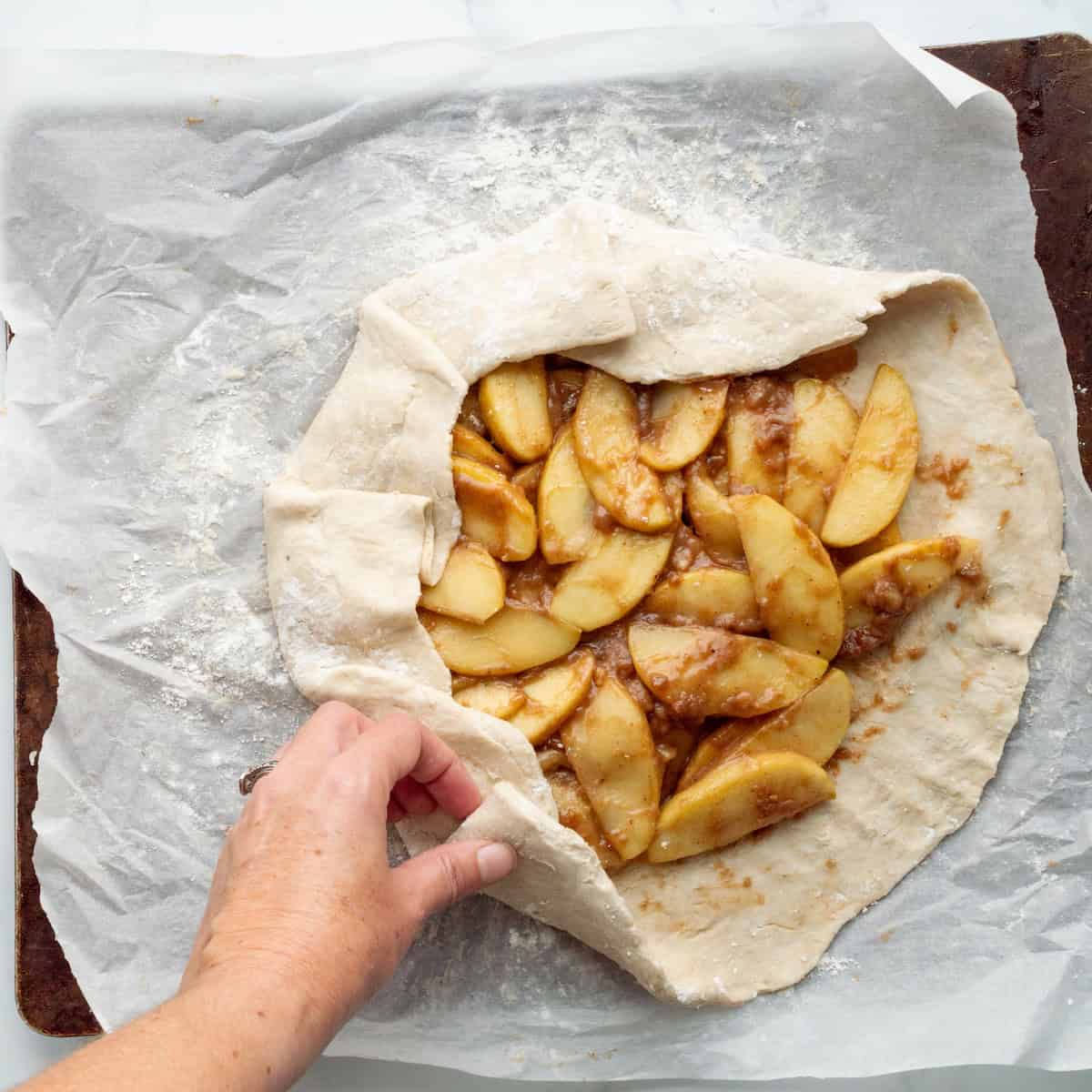 Folding the pie dough over apple filling on a parchment lined baking sheet.