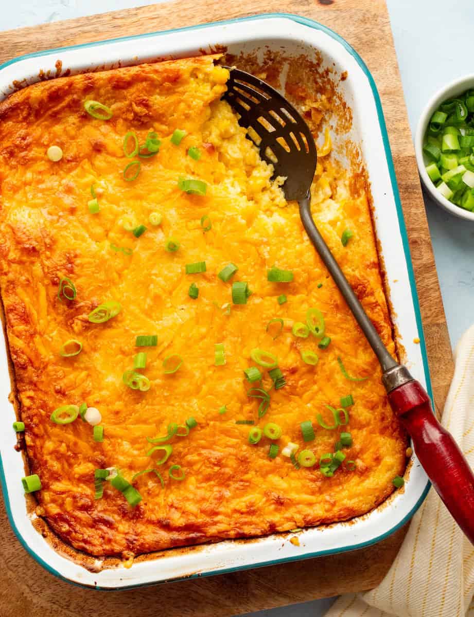 Teal rimmed white enamel pan filled with corn souffle recipe on a wooden board with a red handled spoon and a bowl of green onions and yellow striped cloth on the side.