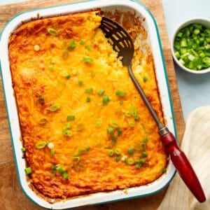 Teal rimmed white enamel pan filled with corn souffle recipe on a wooden board with a red handled spoon and a bowl of green onions and yellow striped cloth on the side.