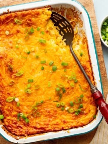 Teal rimmed white enamel pan filled with corn souffle recipe on a wooden board with a red handled spoon and a bowl of green onions and yellow striped cloth on the side.